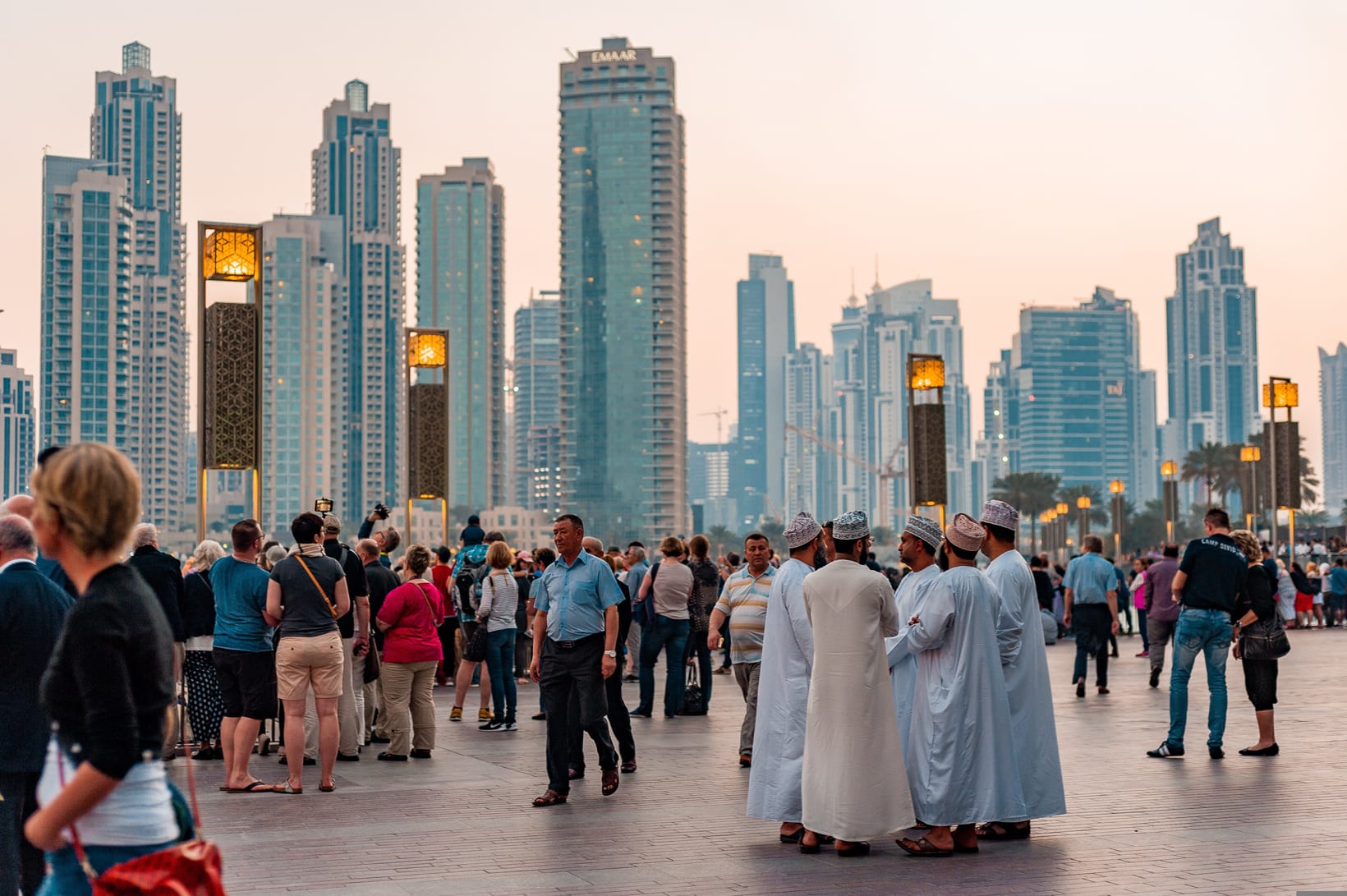 Emirati men in kandura and visitors from many nationalities gathered on the promenade in Downtown Dubai at dusk.