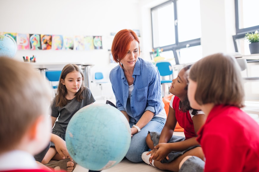 A Teacher sitting on the floor with a group of pupils gathered around a globe in a bright classroom.